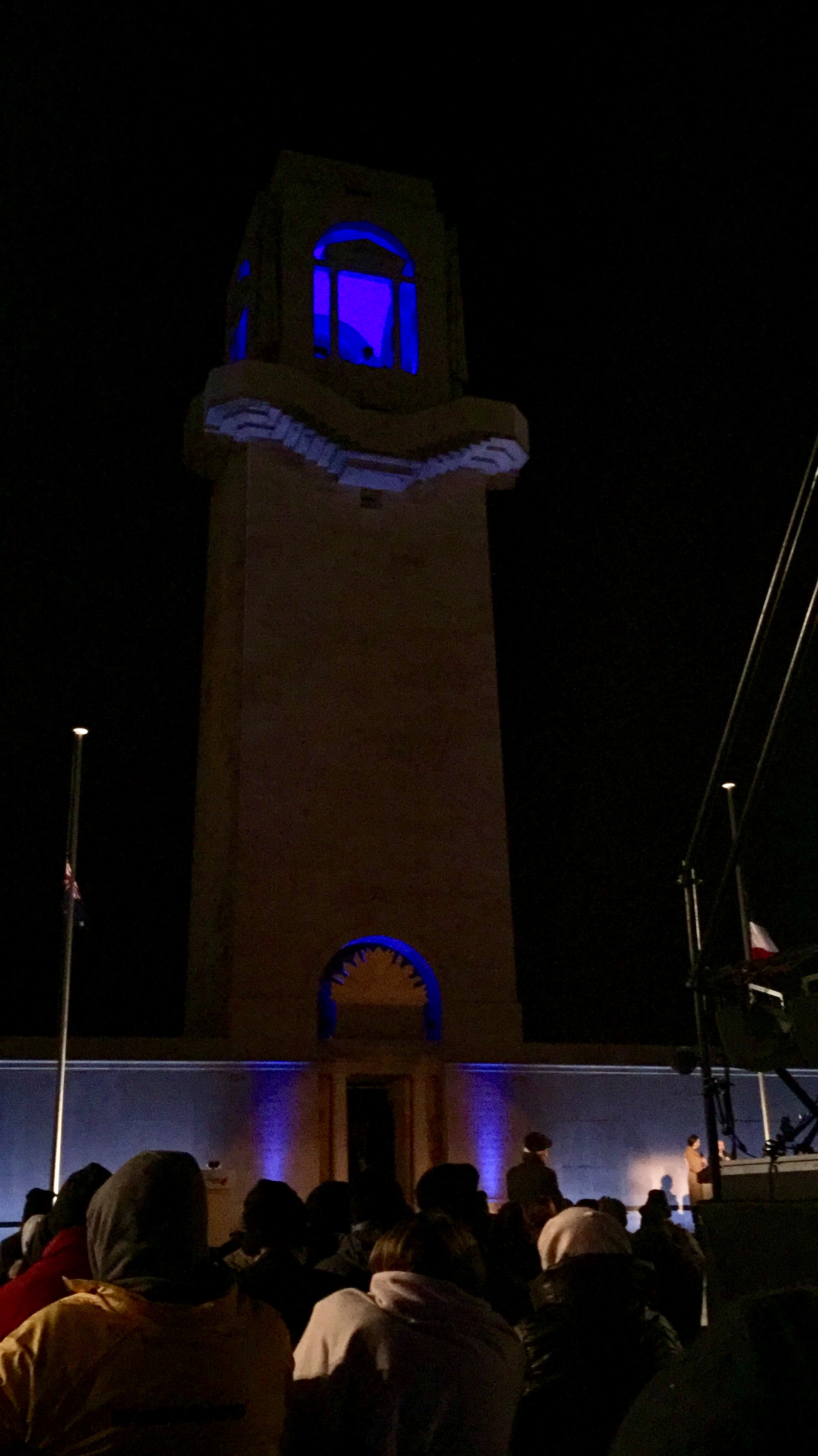 The Australian war memorial at Villers Bretonneux lit up. A crowd is in front and there is a stage with people on it.