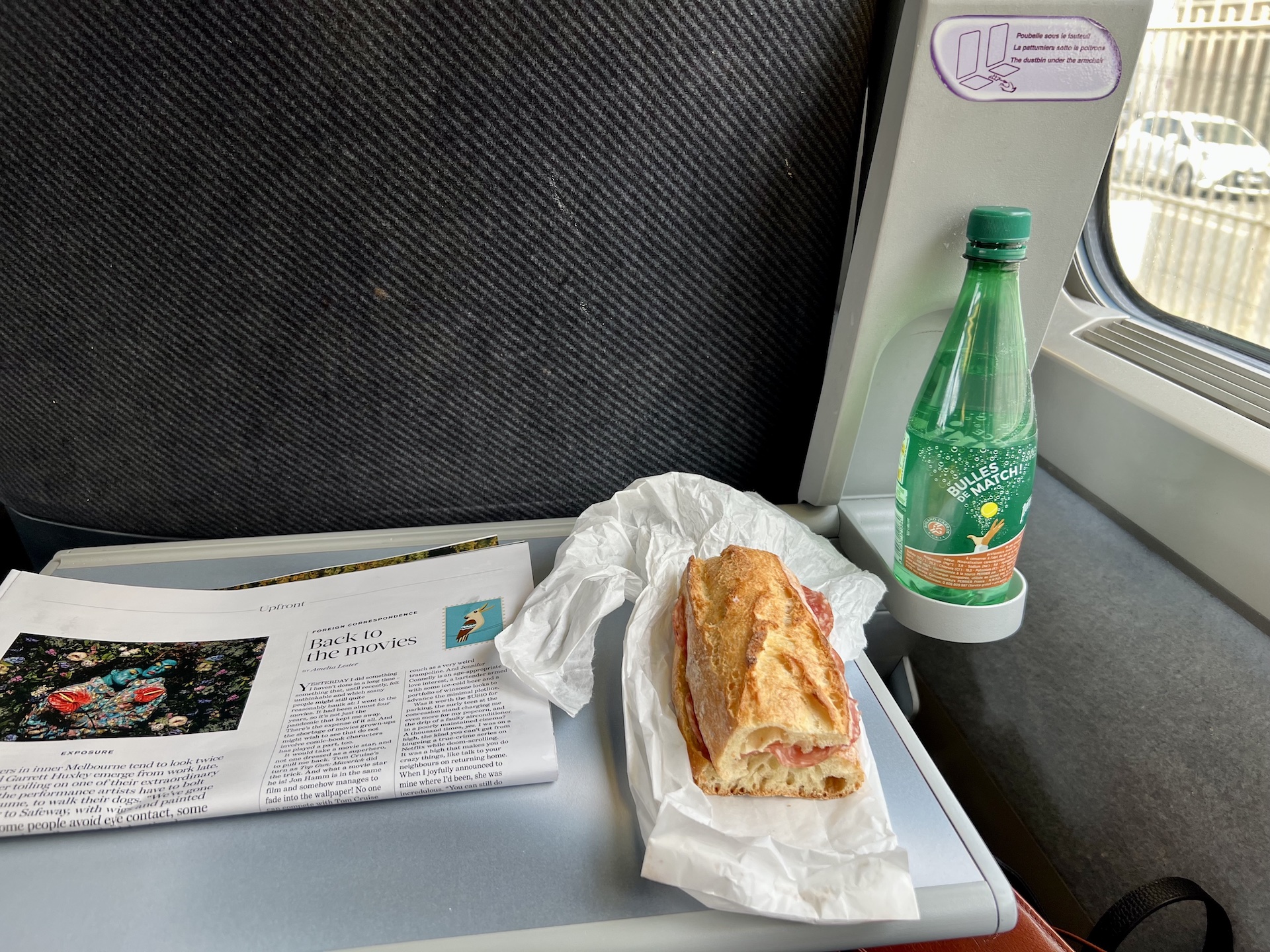 The tray table of a train with a bottle of water, a baguette and a newspaper